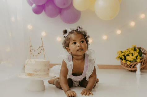 Menina de 1 ano ao lado de um bolo branco e cesta com rosas amarelas e balões na parede