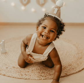 Bebê em ensaio fotográfico de aniversário enquanto está no chão em cima de um tapete com chapéu de aniversário